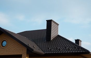 Roof of a brick house with dark shingles and two chimneys against a clear blue sky.